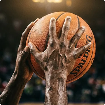 Des mains agrippant fermement un ballon de basket Spalding, prêtes à entrer en action. L'image dégage une impression de concentration et de détermination sur fond de terrain de basket en salle flou.